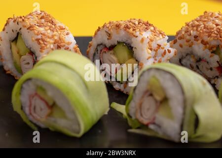Vista in primo piano di un rotolo di sushi californiano con semi di sesamo e surimi in un sushi su un piatto nero e sfondo giallo brillante Foto Stock