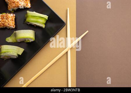 Panini fatti in casa in california e sushi di cetrioli con semi di sesamo su una piastra quadrata nera su sfondo marrone beige con bastoncini di legno Foto Stock