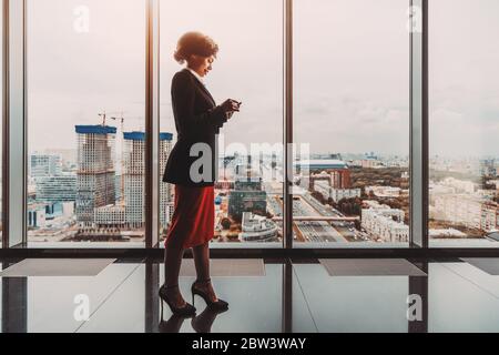 Silhouette di una simpaticissima donna d'affari afro-americana che usa il suo smartphone per rispondere a un messaggio mentre si trova di fronte a una finestra panoramica Foto Stock