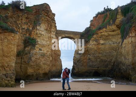 Coppia baciando in Praia dos estudantes spiaggia con arco ponte a Lagos, Portogallo Foto Stock