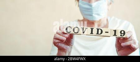 Una donna anziana con una maschera medica sul viso tiene i cubetti con l'iscrizione Covid-19. Il concetto - caronovirus Covid-19. Foto Stock