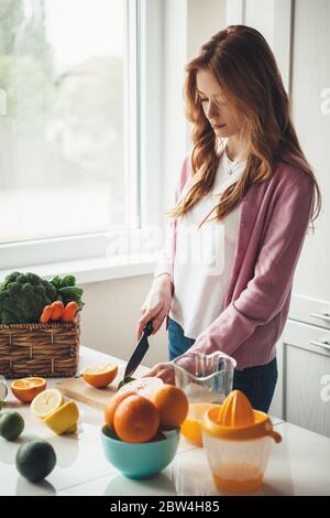 Madre caucasica per il fitness con capelli rossi e fracelle tagliando frutta e facendo succo fresco naturale in cucina da arance e limone Foto Stock