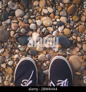 In piedi su Pebbles e Rocks sulla spiaggia guardando giù e la mia vista piedi Blue Shoes Foto Stock