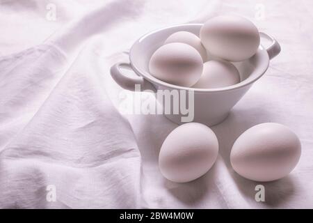 sulla tovaglia di lino bianco, in primo piano, una ciotola in ceramica con uova fresche bianche pronta per la preparazione di ricette culinarie fatte in casa Foto Stock