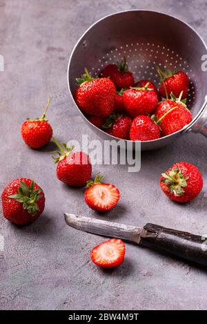 Su uno sfondo grigio testurizzato, fragole mature in un vecchio colander e alcune tagliate con un coltello vintage. Foto Stock