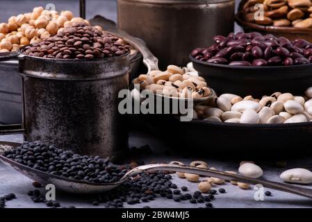 in diverse ciotole di metallo rustico, su uno sfondo grigio testurizzato, varietà di legumi secchi crudi e colorati ricchi di proteine Foto Stock