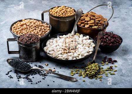 in diverse ciotole di metallo rustico, su uno sfondo grigio testurizzato, varietà di legumi secchi crudi e colorati ricchi di proteine Foto Stock