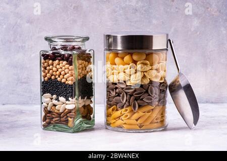 Su uno sfondo grigio testurizzato, varietà di pasta italiana e legumi secchi in vasi di vetro in primo piano. Dieta mediterranea Foto Stock