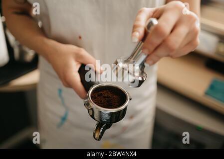 Una foto ravvicinata delle mani femminili che tengono una manomissione metallica e un portafiltro con caffè in una caffetteria. Un barista che si prepara alla spremitura del caffè macinato Foto Stock