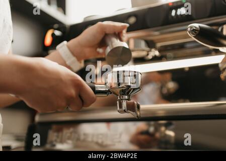 Una foto ravvicinata delle mani femminili che tengono una manomissione metallica e un portafiltro con caffè in una caffetteria. Un barista che si prepara alla spremitura del caffè macinato Foto Stock