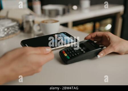 Una donna che paga il suo caffè con uno smartphone con la tecnologia SENZA contatto PAY PASS in un bar. Una barista femminile dispone di un terminale per pagare a un cl Foto Stock