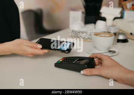 Una ragazza che paga il latte con uno smartphone grazie alla tecnologia NFC senza contatto in un bar. Una barista femminile dispone di un terminale per pagare a un cliente Foto Stock