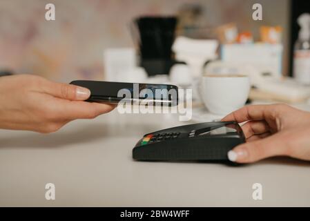 Una donna che si prepara a pagare per il latte con uno smartphone con tecnologia NFC senza contatto in un bar. Una barista femminile dispone di un terminale per pagare Foto Stock