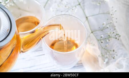 Dalla teiera in vetro trasparente versare il tè dorato in una tazza di vetro. Teiera di vetro che versa il tè nero nella tazza. Tavolo di legno bianco Foto Stock