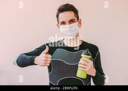 Felice uomo fitness istruttore in maschera medica volto mostrando il pollice in su gesto. Giovane uomo sportivo sicuro in maschera protettiva gesturing come per l'allenamento Foto Stock