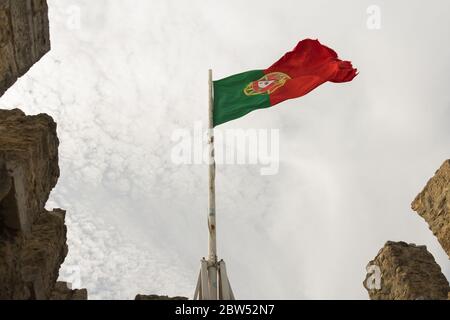 Bandiera del Portogallo (rossa, gialla e verde), Lisbona. Foto a basso angolo del castello di São Jorge nella parte storica della capitale. Nuvoloso e ventoso Foto Stock