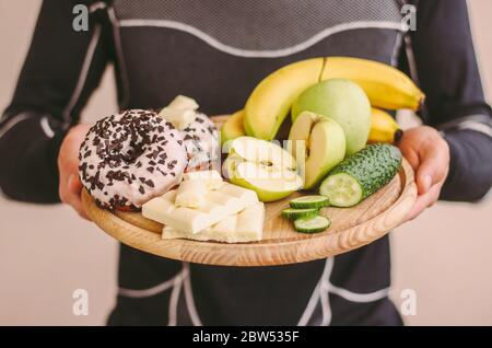 Primo piano di un giovane uomo fitness che decide con la scelta tra frutta sana e zucchero cibo spazzatura malsano. Sport uomo tenere tavola di legno con mele, bana Foto Stock