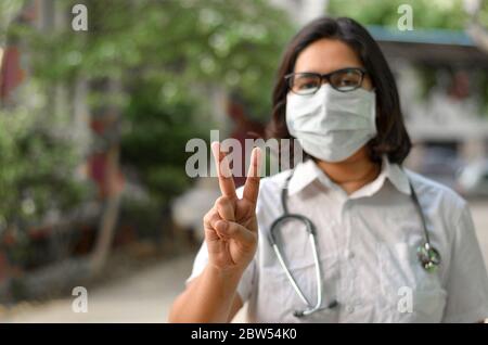 Ritratto di una giovane operatrice medica femminile che fa un segno di vittoria con la mano destra che indossa la maschera chirurgica per proteggersi da Corona VI Foto Stock
