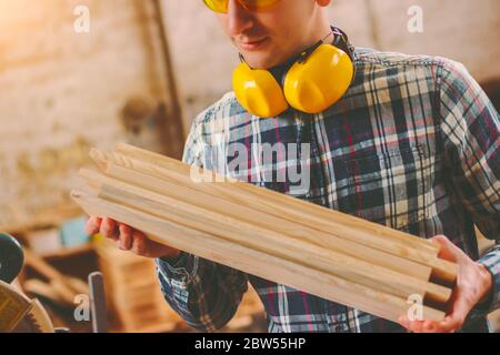 Primo piano di giovane falegname maschile con cuffie protettive sul collo che tiene in mano una pila di tavole di legno mentre si lavora in segheria. arti professionali Foto Stock