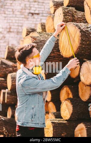 Responsabile magazzino giovane uomo in occhiali protettivi e cuffie per il controllo delle dimensioni del materiale in legno tagliato. Forestiere maschio, carpentiere con nastro di misurazione Foto Stock