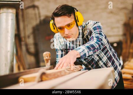 Produttore professionale di armadi con cuffie e occhiali protettivi che lavora con la macchina da taglio in officina per la lavorazione del legno. Falegname maschio con elet Foto Stock