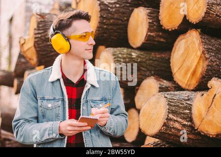 Ritratto di giovane forestere in occhiali protettivi e cuffie controllo albero taglio tronchi inventario al magazzino legname. Falegname lavoratore Countin Foto Stock