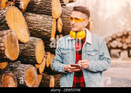 Ritratto di sicuro uomo magazzino legname manager in occhiali protettivi e cuffie contare legno tagliato inventario. Carpentiere giovane che controlla cu Foto Stock