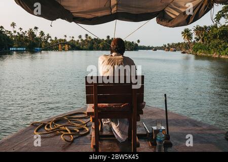 L'autista di una casa galleggiante Keralan guida la barca da un sedile sulla prua, come il sole tramonta Foto Stock