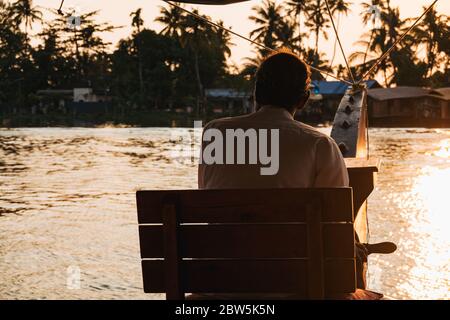 L'autista di una casa galleggiante Keralan guida la barca da un sedile sulla prua, come il sole tramonta Foto Stock