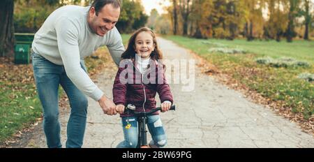 Un padre attento insegna a sua figlia come guidare la bicicletta aiutandola nel parco sorridendo Foto Stock