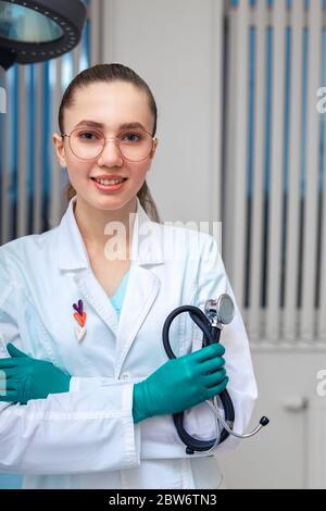 Giovane donna dottore in un cappotto con un fonendoscopio in mani che si pone alla telecamera sullo sfondo del reparto in ospedale. Copia spazio, Medecine Foto Stock