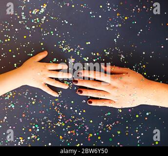 Vista dall'alto della pianta anonima con manicure e bambino che tiene le mani sulla superficie nera cosparsa di colorati confetti Foto Stock