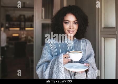 Sorridi afroamericana ragazza in piedi con una tazza di caffè avvolto in plaid. Bella signora con capelli scuri ricci bere caffè nel ristorante Foto Stock