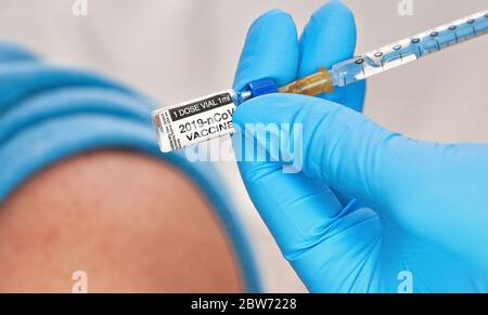 Indossare guanti blu contenenti siringa e vaccino del coronavirus (disegno di un adesivo, dati fittizi), in procinto di iniettarlo alla spalla del paziente seduto vicino, Foto Stock