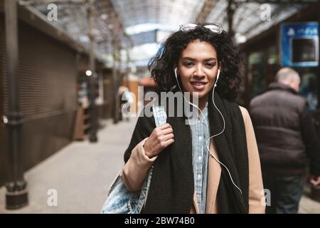 Ritratto di bella ragazza afroamericana in piedi sulla strada e guardando in macchina fotografica Cool ragazza con capelli ricci scuri in auricolari che girano per la città Foto Stock