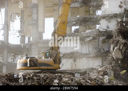 Primo piano di un escavatore idraulico-distruttore speciale durante il funzionamento, polvere di calcestruzzo nell'aria, rovine di un edificio sullo sfondo. Demolizione ultra elevata Foto Stock