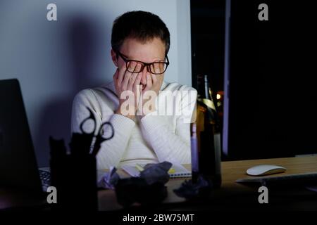 Un giovane esausto in occhiali che sfrega gli occhi stanchi lavorando a tarda notte, bevve una birra per rilassarsi, si addormenta dalla fatica. Lavoro eccessivo, pigrizia, occhi Foto Stock