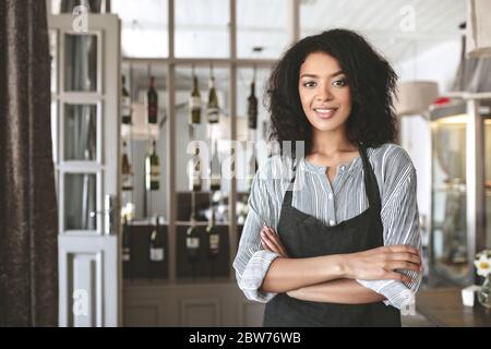 Giovane afroamericana in grembiule in piedi con le braccia ripiegate nel ristorante. Bella ragazza con capelli scuri ricci in piedi in grembiule al caffè Foto Stock