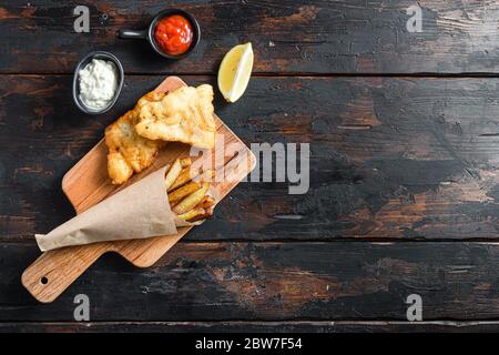 Pesce e patatine in un cono di carta su legno tritare bordo tuffo e limone - merluzzo fritto, patatine fritte, fette di limone, salsa tartaro, ketchup tomatoe con mu Foto Stock