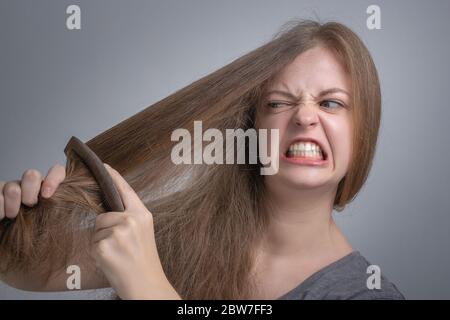 Giovane donna caucasica ragazza che lotta per pettinare i suoi capelli diritti con il viso divertente frustrata. Concetto di problema dei capelli lunghi Foto Stock