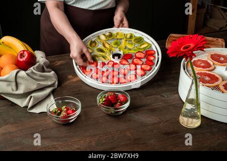 Mani di donna di mezza età che mettono fragole e kiwi a fette su vassoio di frutta asciuga tra banane fresche, arance, ecc su tavolo da cucina Foto Stock