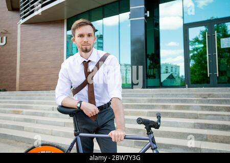 Felice giovane uomo d'affari riuscito che ti guarda mentre si appoggia sulla sua bicicletta sullo sfondo di edificio esterno e scala Foto Stock