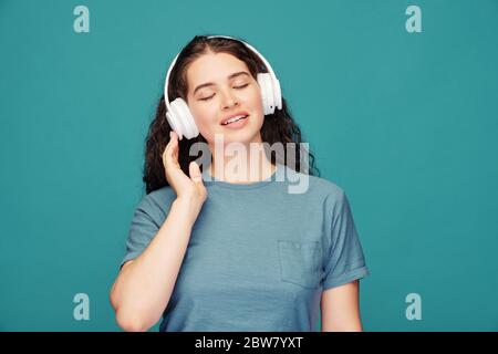 Felice giovane donna bruna in t-shirt blu e cuffie bianche che ascolta musica rilassante mentre si trova davanti alla fotocamera Foto Stock