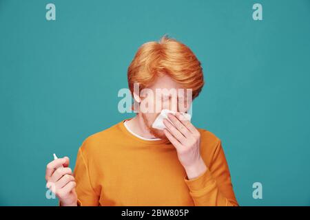 Giovane allergico che tiene il fazzoletto di carta dal naso mentre starnutisce e usando lo spray antiallergico davanti alla macchina fotografica in isolamento Foto Stock