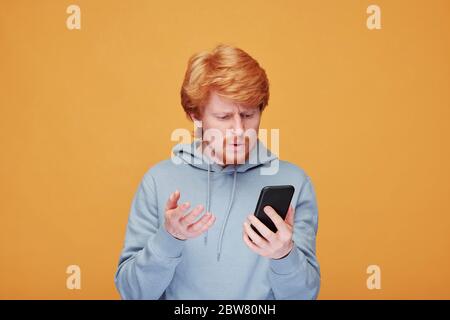 Giovane uomo confuso o insoddisfatto con cappuccio che parla con qualcuno mentre si guarda lo schermo dello smartphone su sfondo giallo Foto Stock