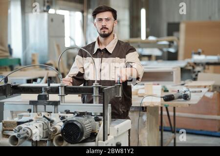 Giovane lavoratore serio di fabbrica di mobili che guarda mentre si sta in piedi da banco di lavoro sullo sfondo dell'interno dell'officina durante il lavoro Foto Stock