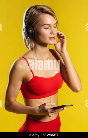 È tempo di ascoltare la musica. Vista laterale di una donna bionda sportiva con cuffie che tengono gli occhi chiusi e ascolta la musica mentre si alza contro il giallo Foto Stock