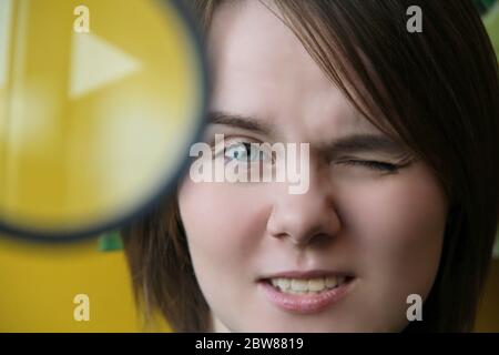 Una bella ragazza giovane guarda attraverso una lente di ingrandimento che squinting un occhio. Primo piano con il lato di stampa rivolto verso l'alto Foto Stock