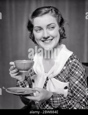 1930 DONNA BRUNA SORRIDENTE CHE INDOSSA ABITO PLAID CON COLLETTO BIANCO CHE TIENE LA TAZZA DI CAFFÈ GUARDANDO FUORI AL LATO - F6807 HAR001 HARS TESTA E SPALLE BEVANDA ALLEGRA SORRISI FLUIDI CONSUMARE IDRATAZIONE GIOIOSA ELEGANTE CAFFEINA JOE JAVA RINFRESCANTE GIOVANE DONNA ADULTA BEVANDE NERO E BIANCO ETNIA CAUCASICA HAR001 VECCHIO ALLA MODA Foto Stock