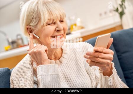 Donna grigia con auricolari che utilizza uno smartphone moderno, trascorrendo la giornata a casa Foto Stock